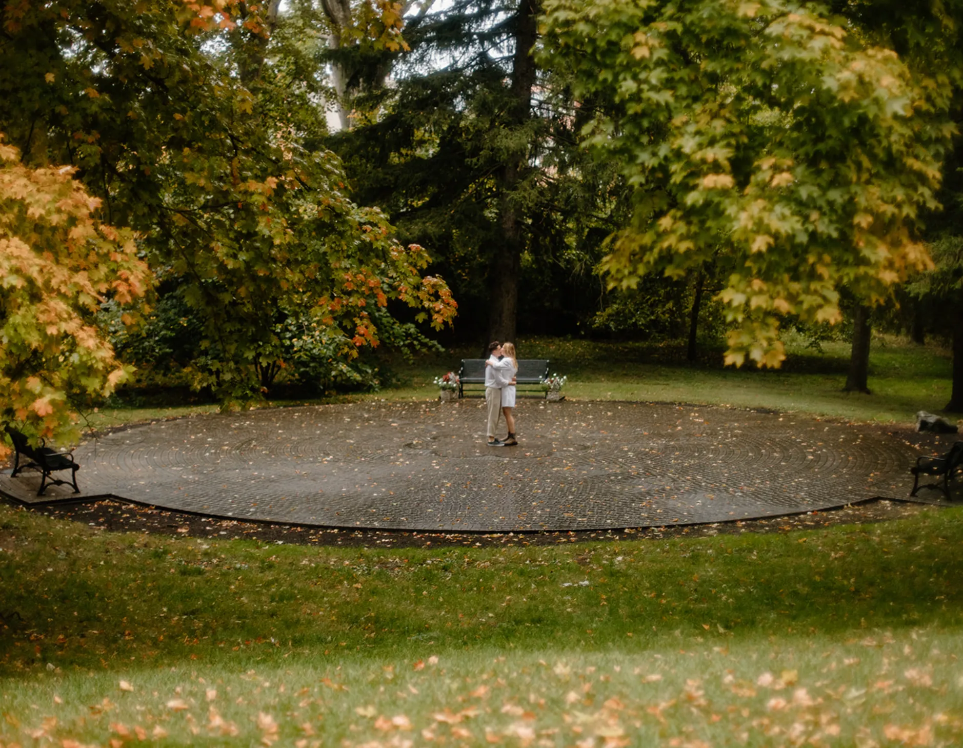 Wide view of the couple embracing in a circular brick plaza under fall trees